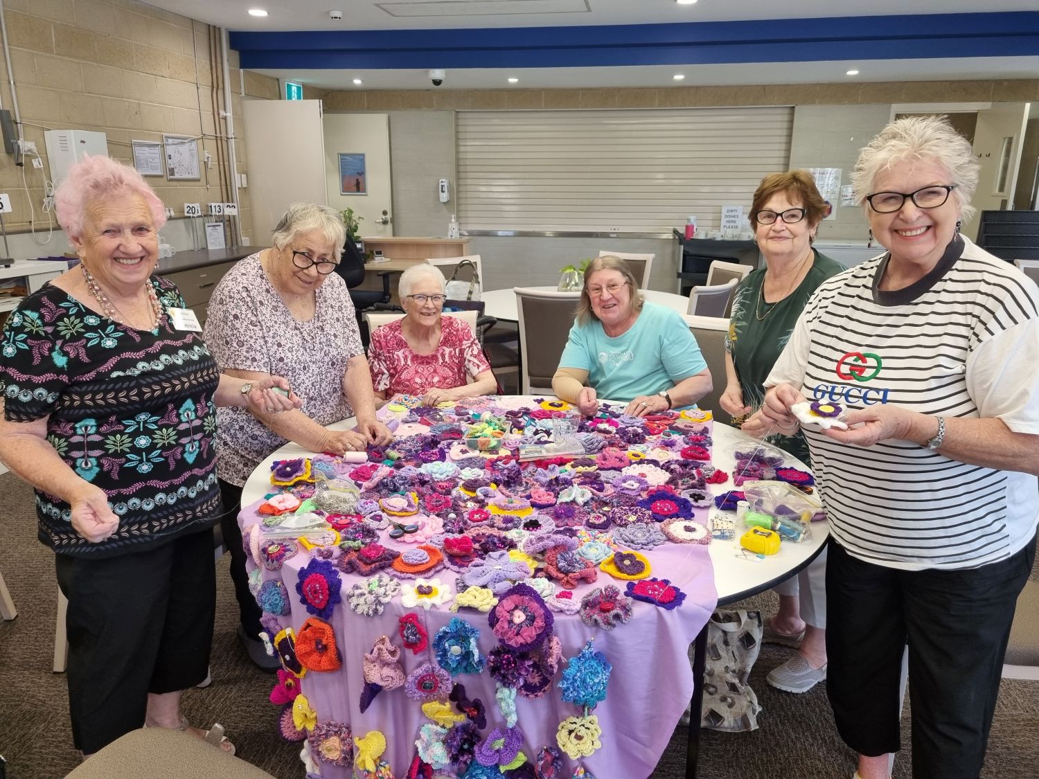Six smiling senior women gather around a large round table covered in hundreds of handmade crochet and knit flowers during a community craft session.